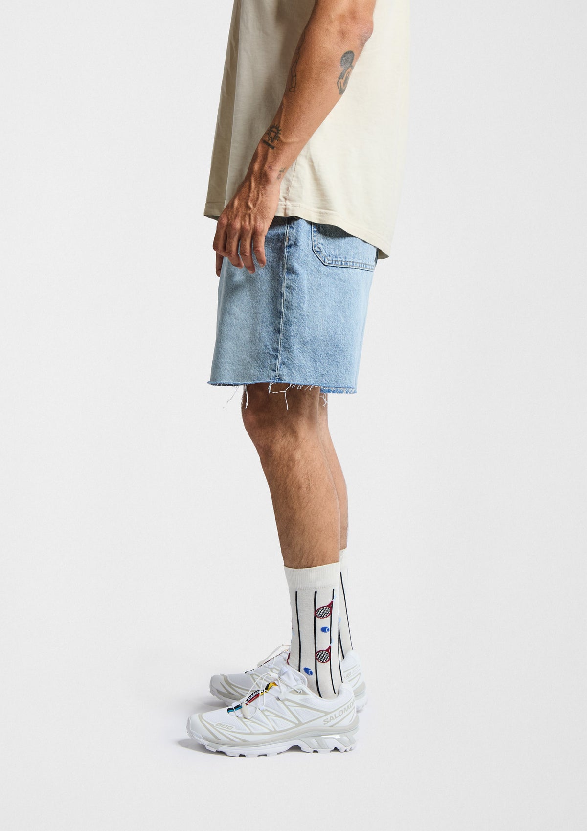 Hombre usando calcetines originales BeTennis con dibujos de raquetas y pelotas de tenis, shorts de mezclilla y zapatillas.